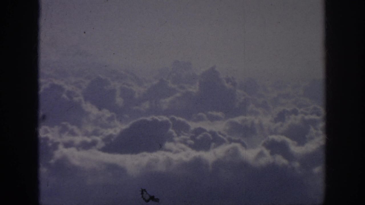 1962 8mm home-movie frame showing a cloud layer from an airplane-window viewpoint, with dark edge shadows and visible film wear.