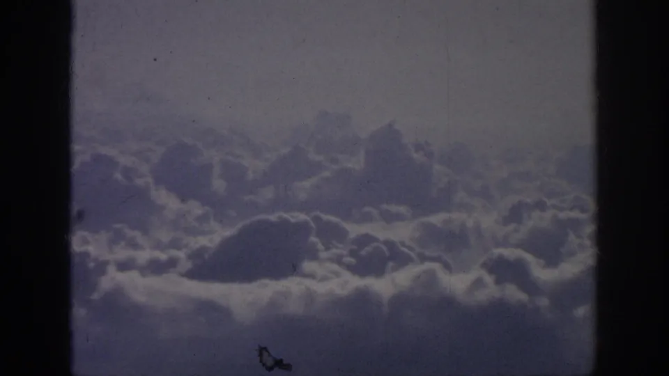 1962 8mm home-movie frame showing a cloud layer from an airplane-window viewpoint, with dark edge shadows and visible film wear.