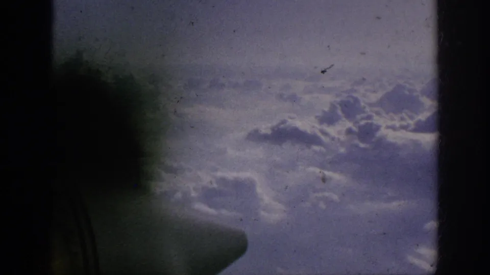 Airplane-window view over clouds in 1962 8mm footage, framed by dark gate edges and a shadowed window shape.