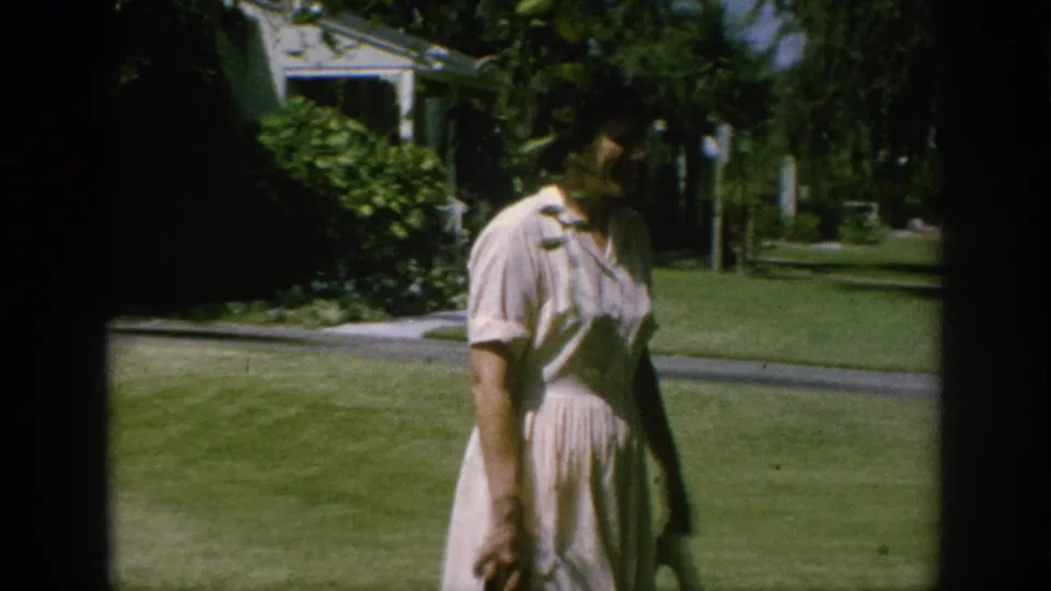 1962 Anaheim home-movie frame of a woman in a light dress walking on a suburban lawn with shrubs, trees, and a house behind her.