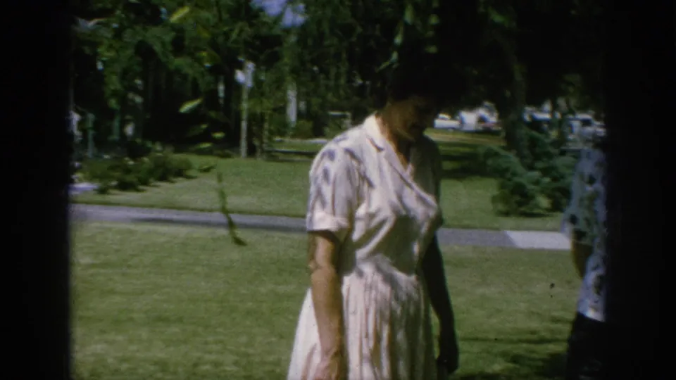Woman in a pale dress crosses a green lawn in Anaheim, with trees, sidewalk, shrubs, and partial figures at the frame edge.
