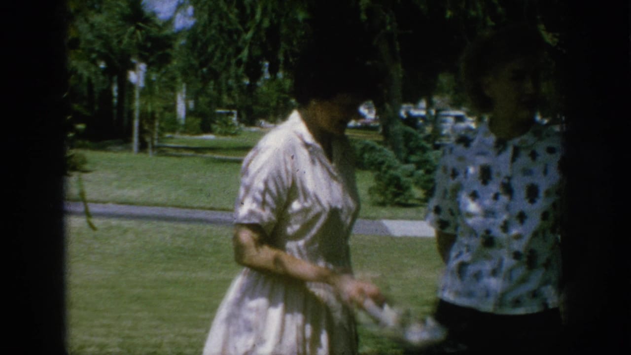Two women stand on a grassy lawn as one holds light-colored high-heeled shoes.