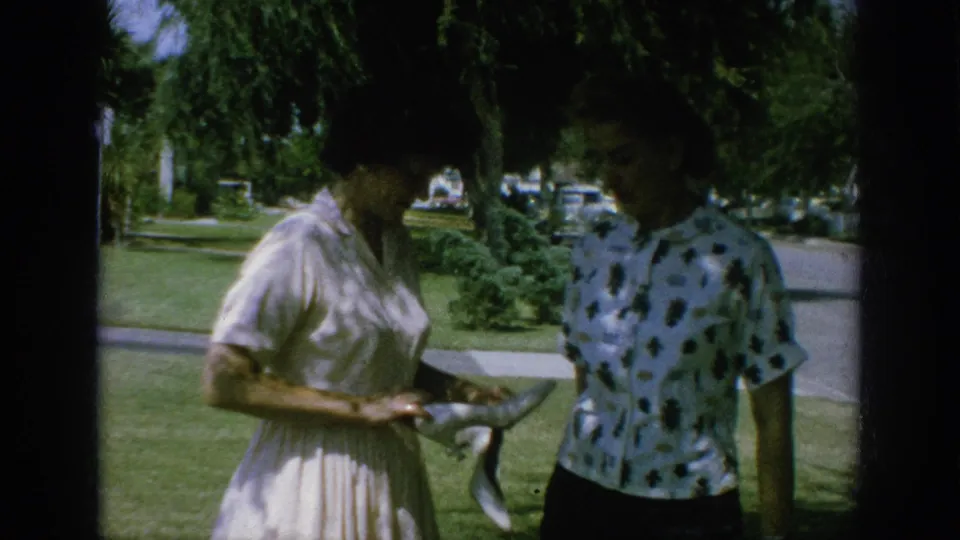 In the 1962 Anaheim home-movie frame, a woman in a light dress presents high-heeled shoes to a woman in a patterned blouse.