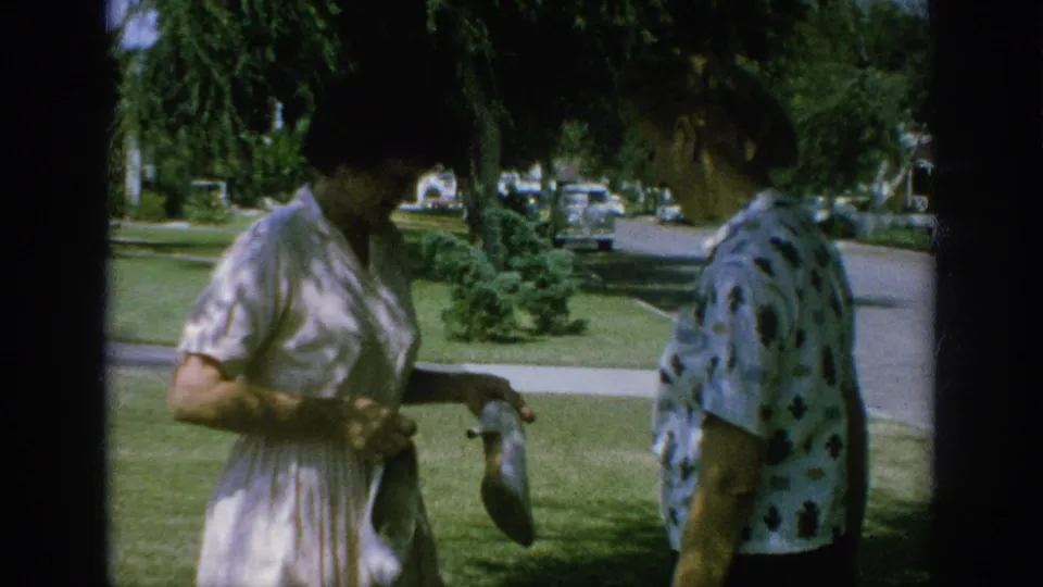 In this 1962 Anaheim frame, a woman in a light dress raises a high-heeled shoe as her companion watches beside a suburban street.
