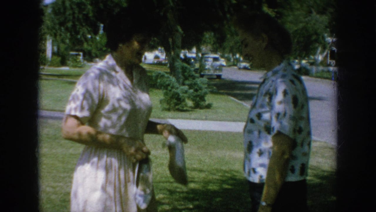 Two women stand on a shaded lawn beside a residential street with parked cars behind them.