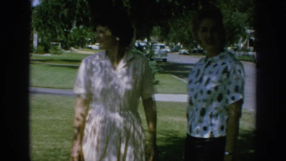 Two adult women in a 1962 Anaheim neighborhood stand under tree shade on a lawn, with mid-century cars visible along the street.