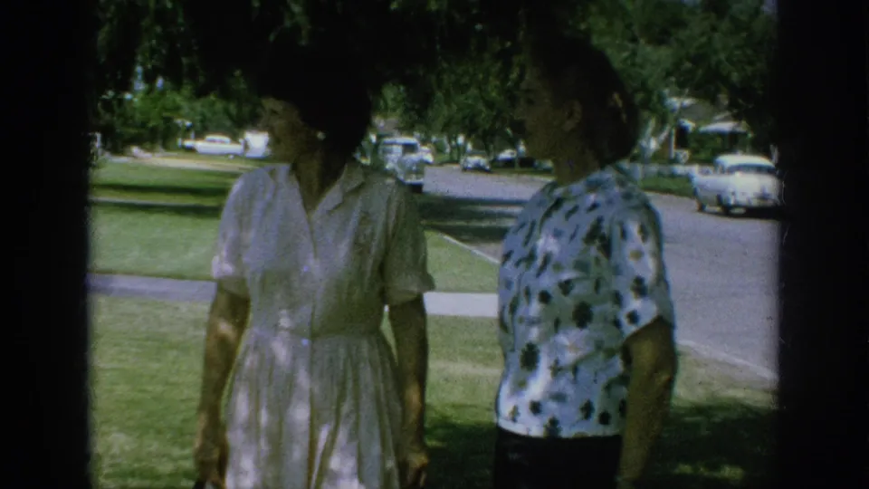 Two adult women in 1962 Anaheim appear to talk on a grassy lawn, with parked cars and a quiet suburban street in the background.