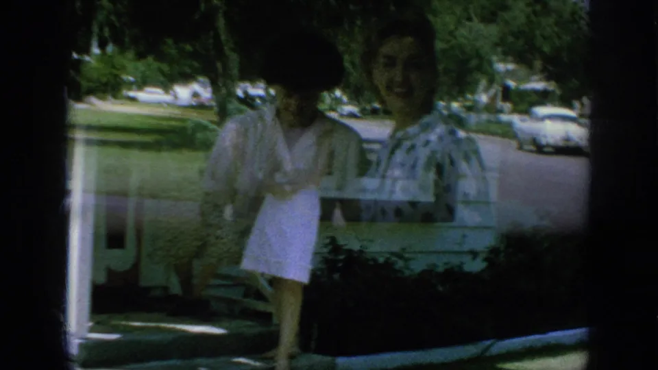 Two adult women in 1962 Anaheim stand near a house exterior with white siding, a window, porch details, and shrubs in front.