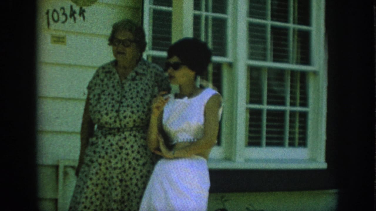 Two women stand closer to a house window, one wearing sunglasses.