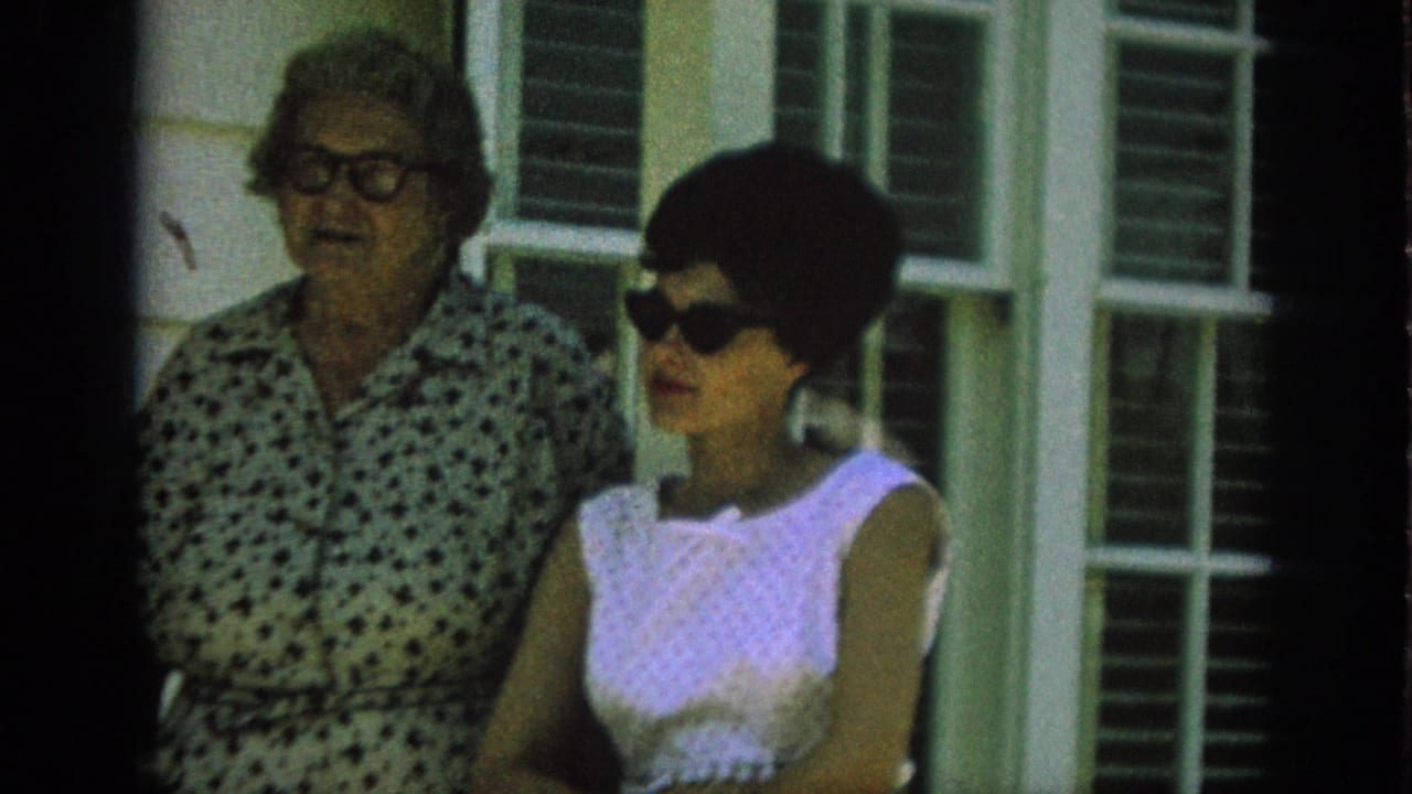 In 1962 Anaheim home-movie footage, two adult women stand near shuttered windows, with one in a white sleeveless dress and dark sunglasses.