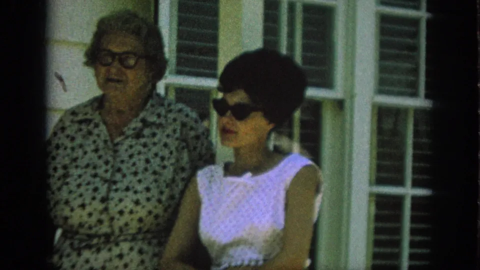 In 1962 Anaheim home-movie footage, two adult women stand near shuttered windows, with one in a white sleeveless dress and dark sunglasses.