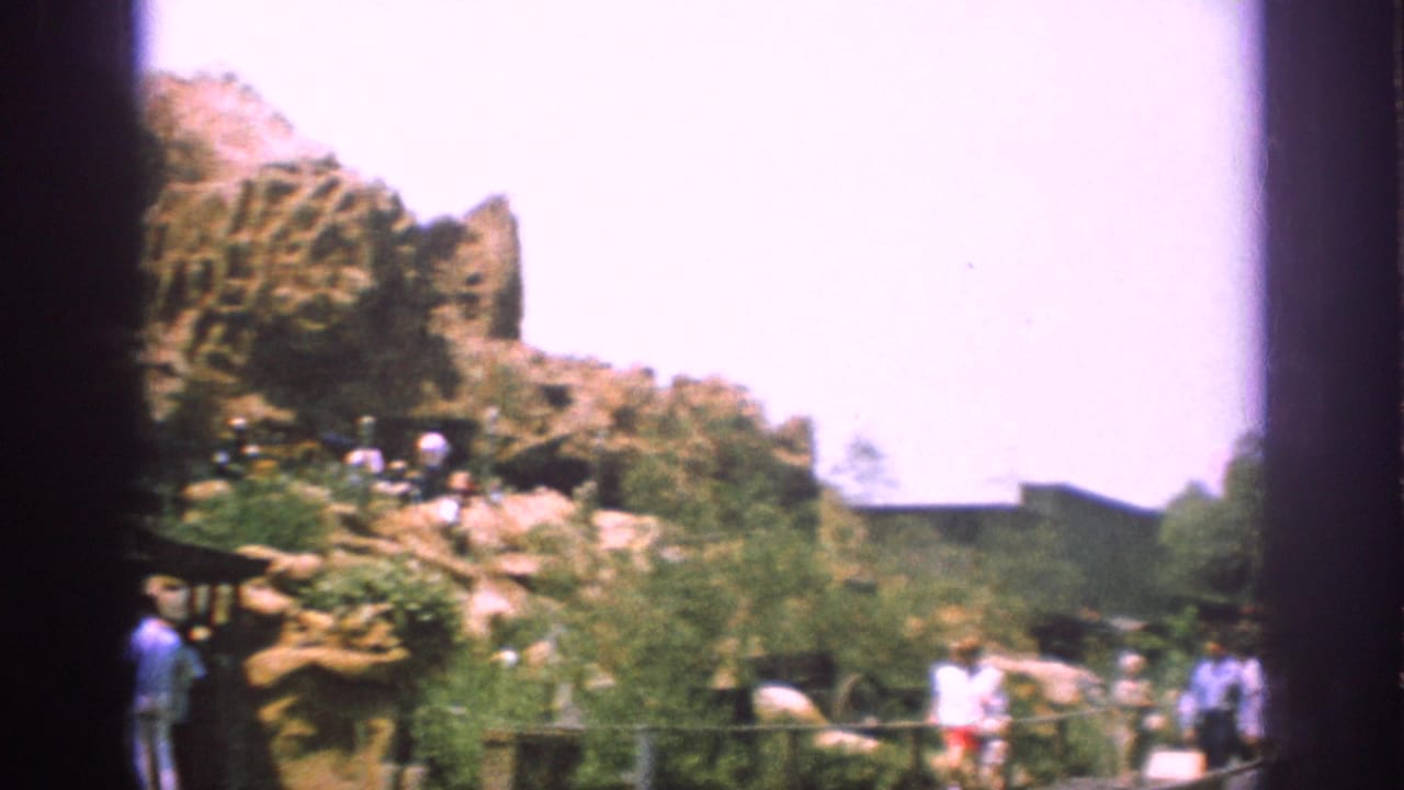 Visitors walk near landscaped artificial rock formations, greenery, railings, and a bright sky.