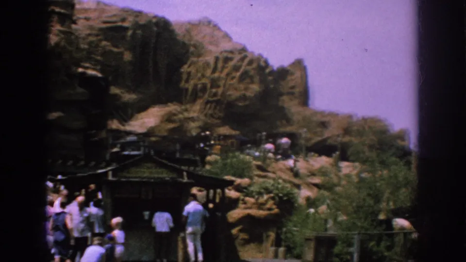 A handheld 1962 Anaheim view shows themed rockwork, plantings, a small roofed structure, and visitors moving along park paths.