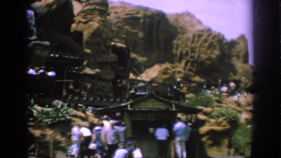 The 1962 Anaheim home-movie frame shows visitors near railings below large artificial rockwork, with greenery and dark film-edge borders.