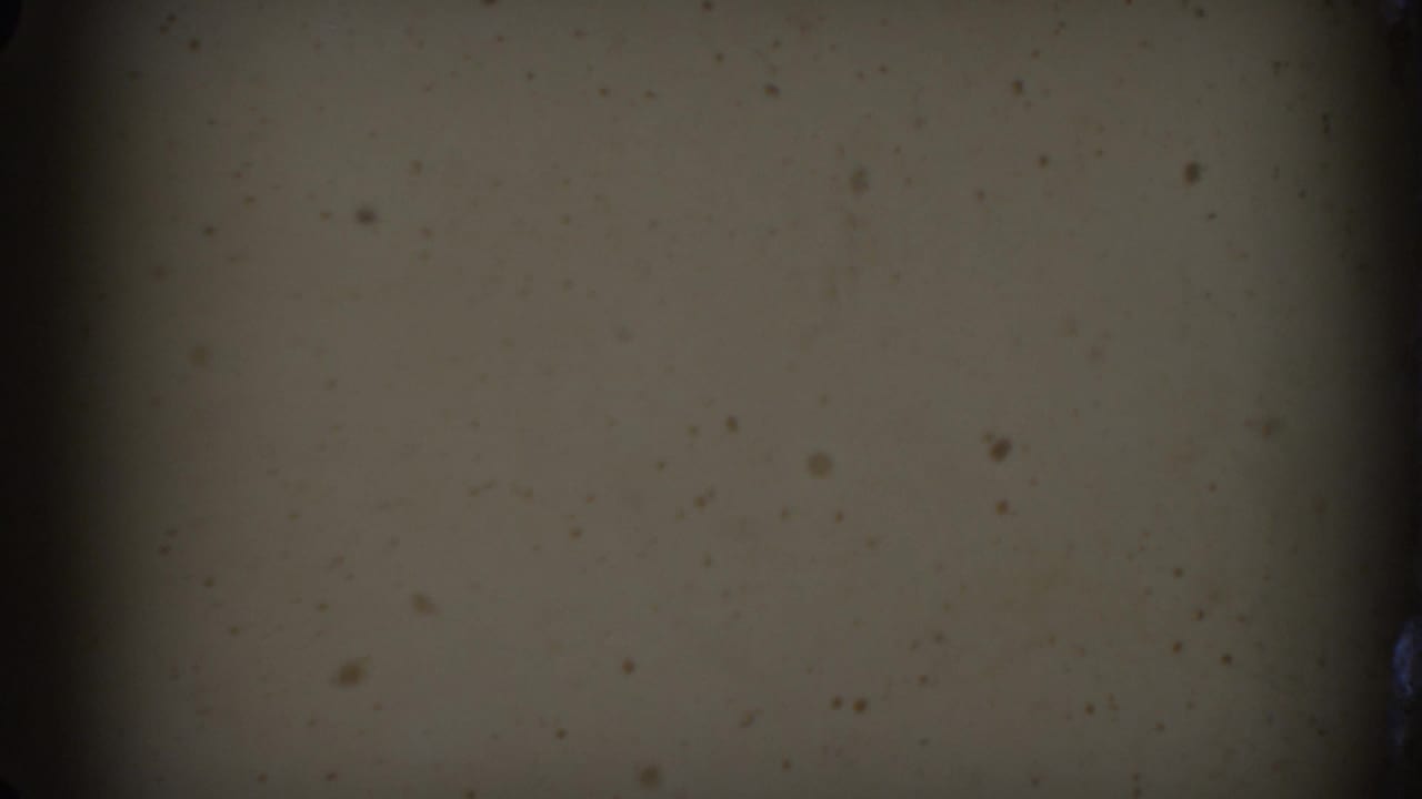 Opening 8mm frame with dark exposure, muted beige surface, black edge areas, and visible film dust before the Raleigh car scene appears.