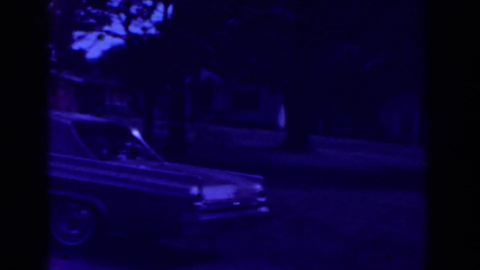 Purple-shifted Raleigh home-movie frame showing part of a long sedan on a residential street with trees and houses in the background.