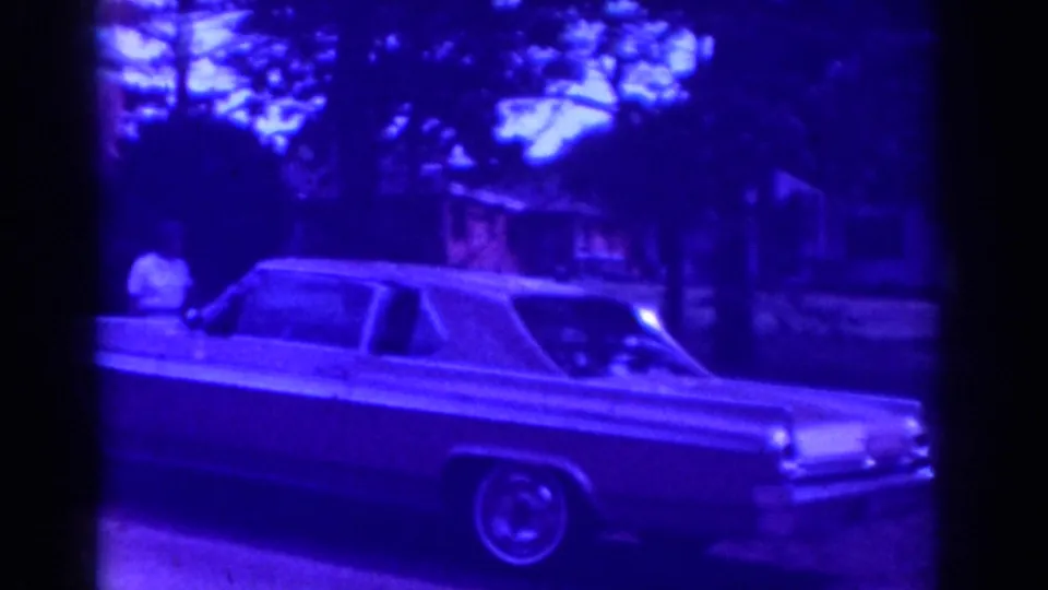 1962 Raleigh frame showing a man in light clothing beside a long mid-century sedan with a whitewall tire in a residential neighborhood.