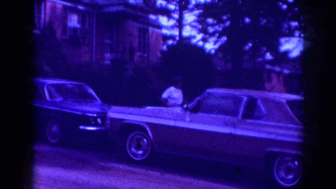 Woman in a white shirt stands near two parked cars on a blue-tinted residential street.