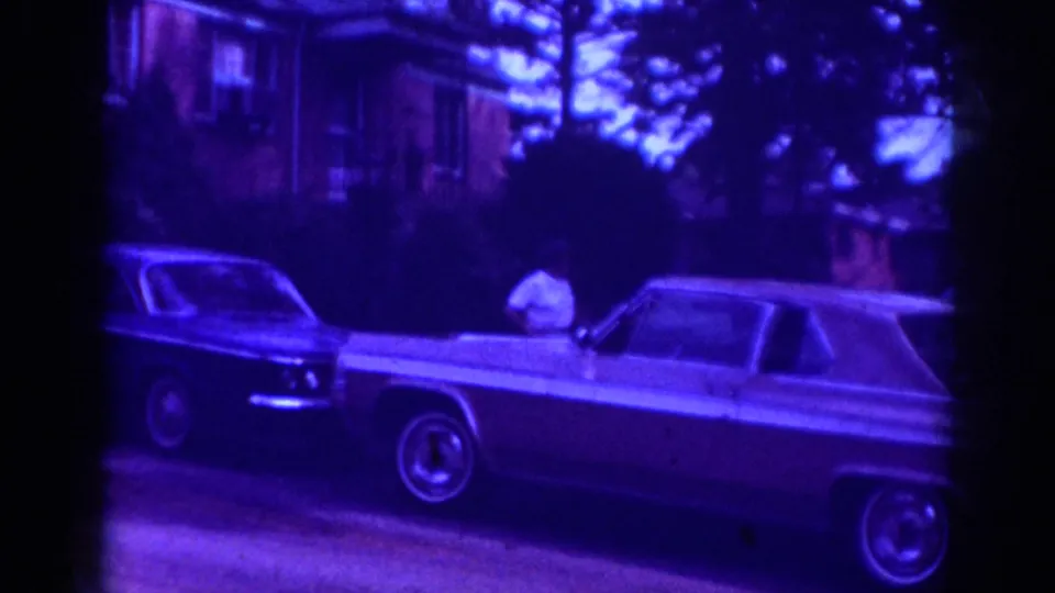 Raleigh 1962 frame showing a woman near a classic sedan and another parked car, with houses and trees behind her.