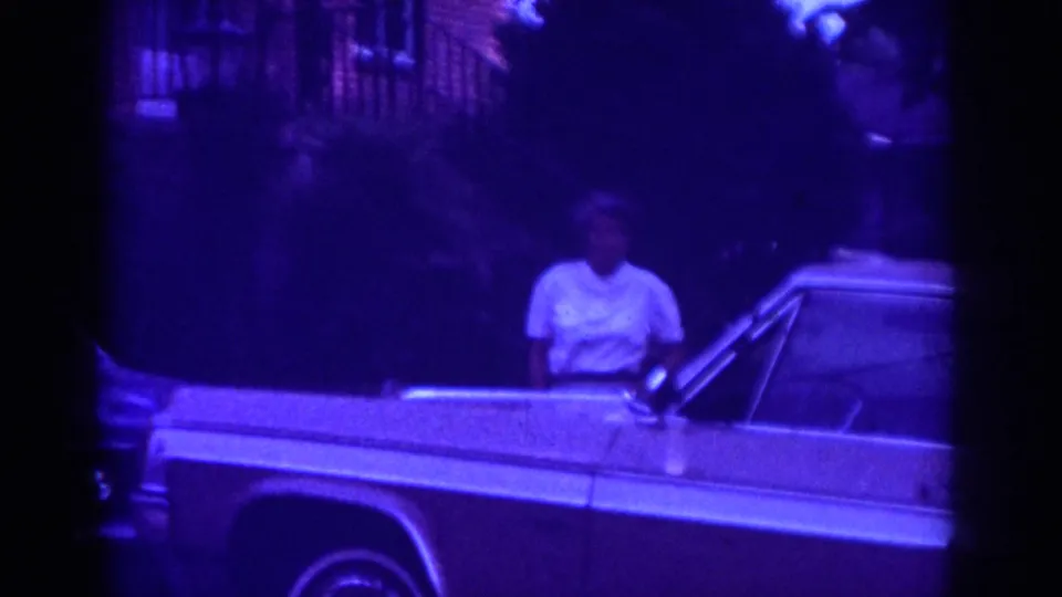 Raleigh 1962 frame showing a woman beside the front of a classic American sedan on a shaded residential street.
