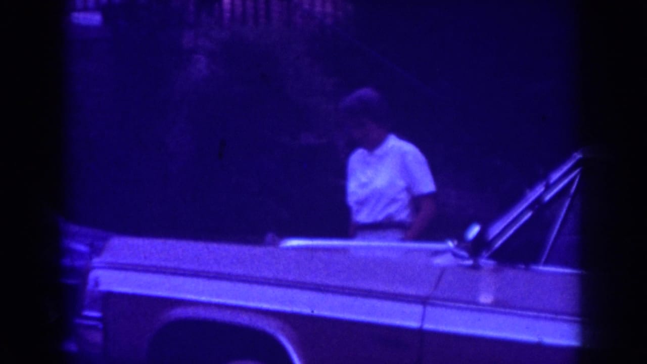 In 1962 Raleigh home-movie footage, a woman in a white top stands near the side of a light-colored classic car at night.