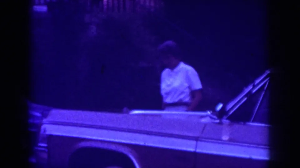 In 1962 Raleigh home-movie footage, a woman in a white top stands near the side of a light-colored classic car at night.