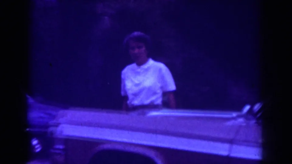 A woman in a white top appears behind a light-colored classic car in nighttime 1962 Raleigh home-movie footage.