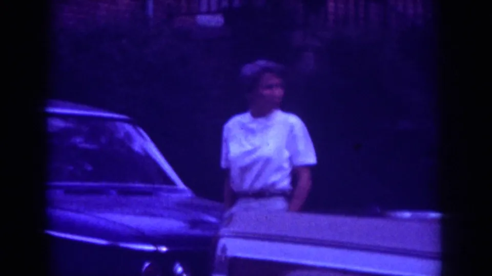 The woman stands near a classic car at night, with another car edge visible in soft 1962 Raleigh 8mm footage.