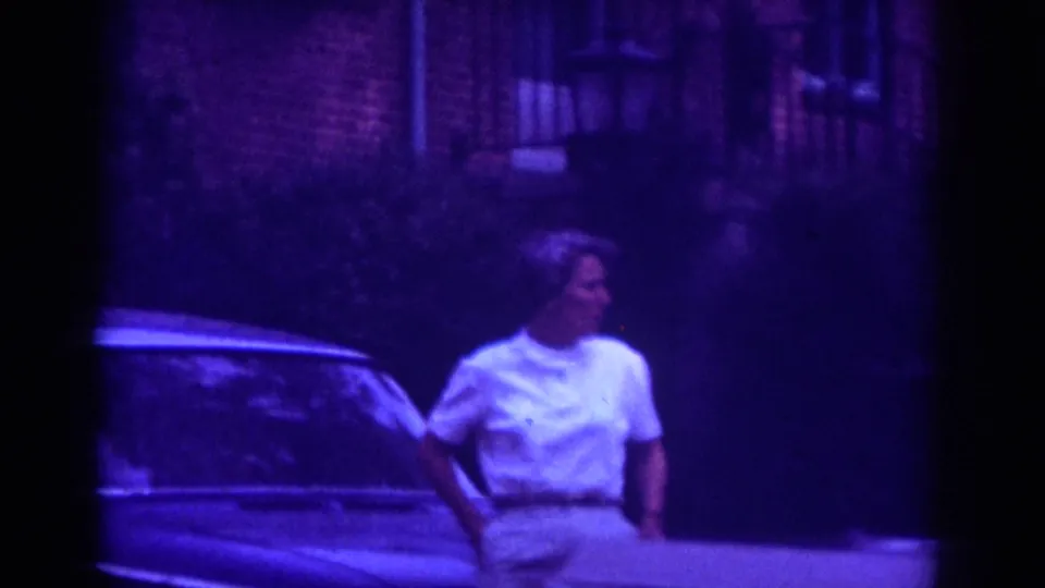 A woman in a white top stands with one arm near a car in purple-blue 1962 Raleigh nighttime home-movie footage.