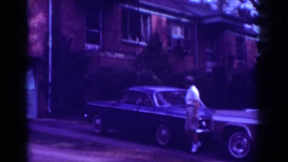 Wider 8mm view shows the woman walking beside a classic car along a residential street in the Raleigh 1962 clip.