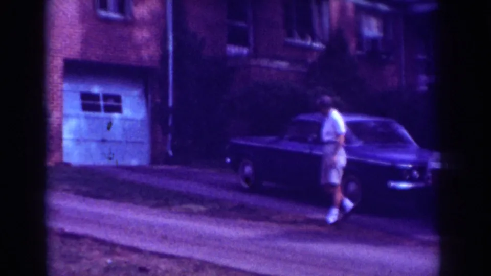 Adult woman in shorts walks past a parked car near a driveway and garage door in a residential Raleigh street scene.