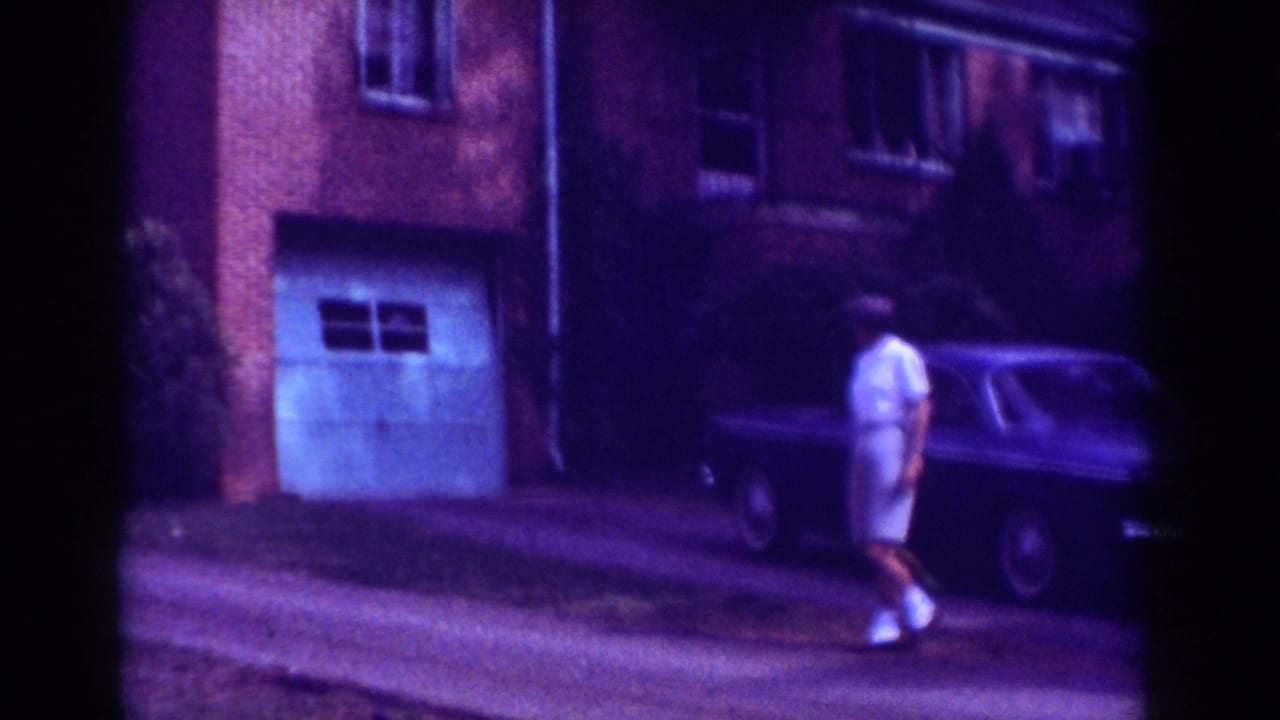 In Raleigh home-movie footage from 1962, a man walks along a driveway beside a brick apartment building and dark parked sedan.