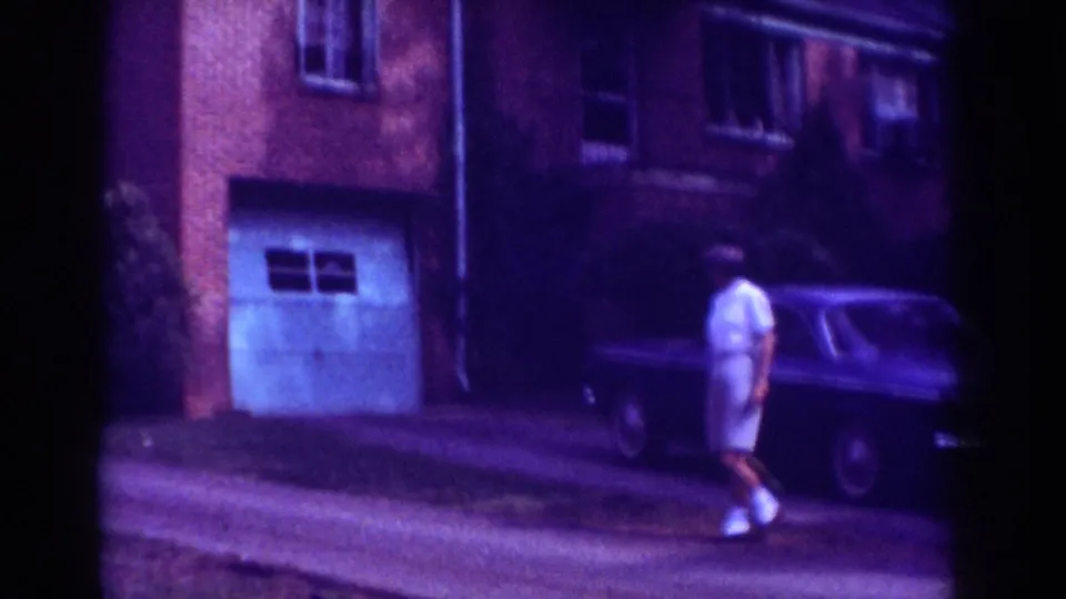 In Raleigh home-movie footage from 1962, a man walks along a driveway beside a brick apartment building and dark parked sedan.