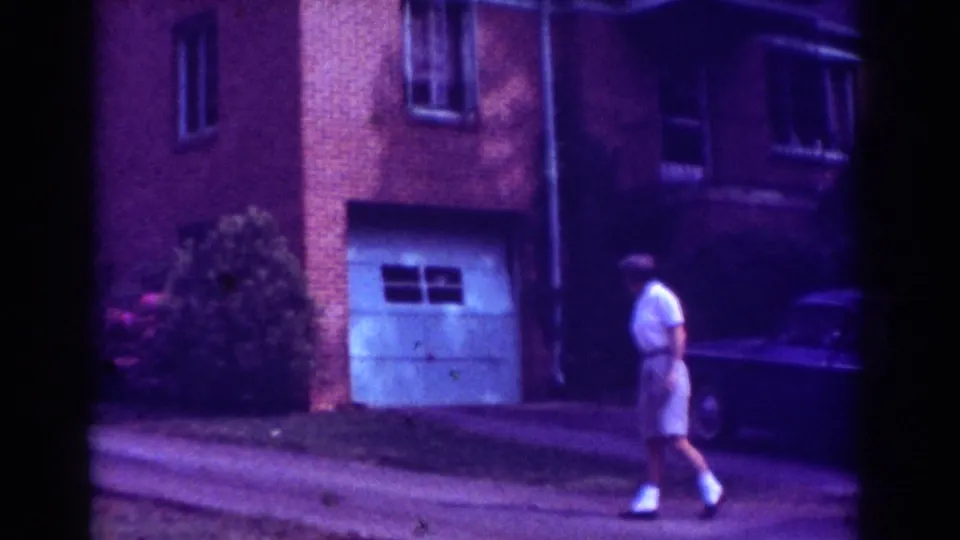 The man continues walking across the residential exterior, with the white garage door, brick wall, shrubs, and parked sedan visible.