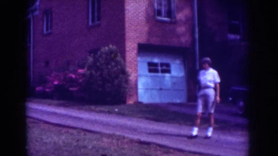 The 1962 Raleigh frame shows the man near the foreground driveway, with shrubs, brick walls, garage door, and part of the parked car behind him.