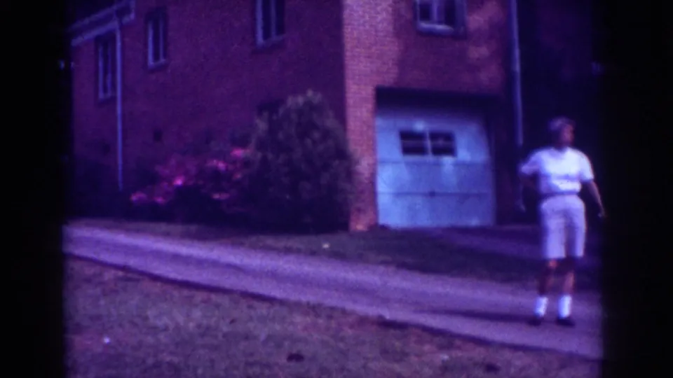 At the end of the clip, the man is near the right edge of the Raleigh residential scene, with the driveway, shrubs, and garage door behind him.