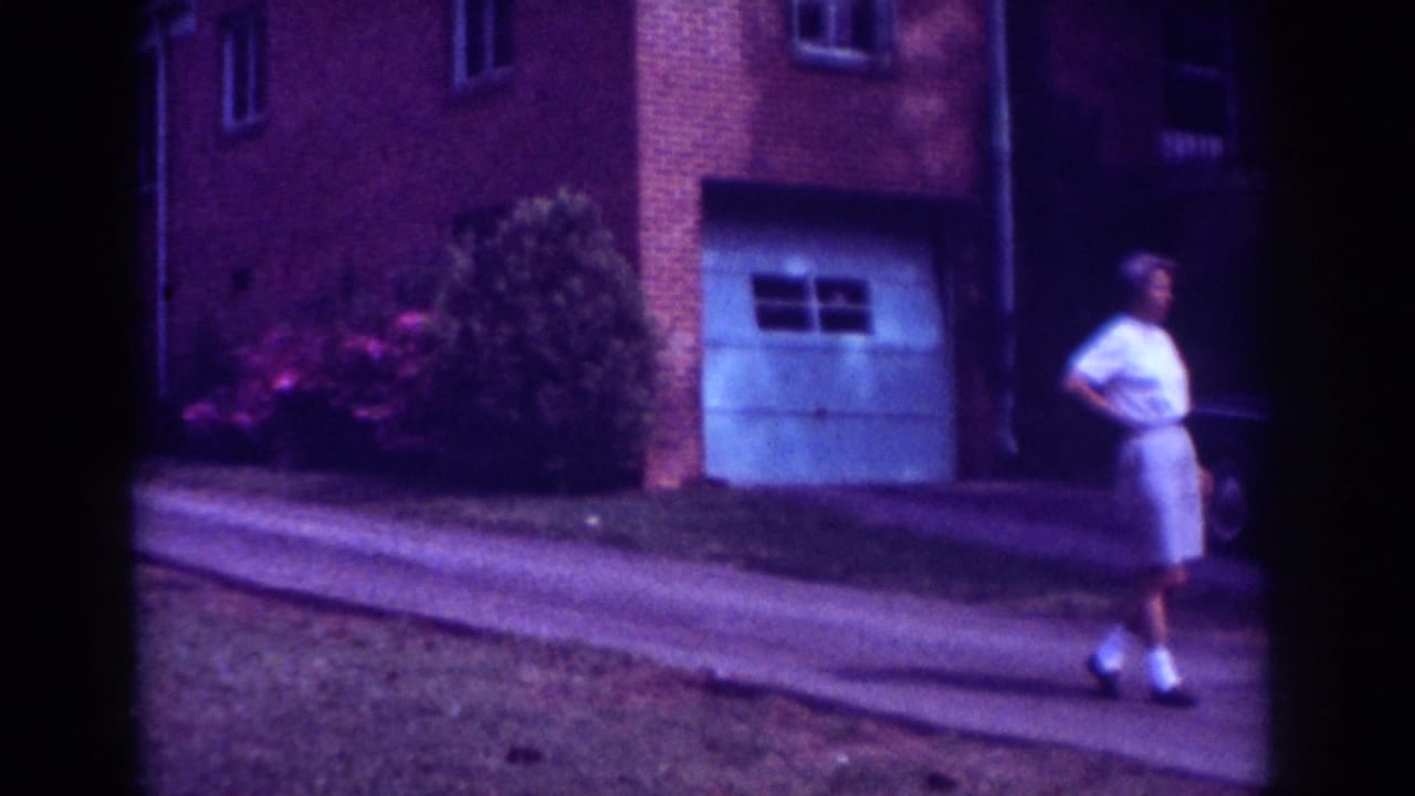 1962 Raleigh home-movie frame showing a person walking down a driveway in front of a brick suburban house with shrubs.