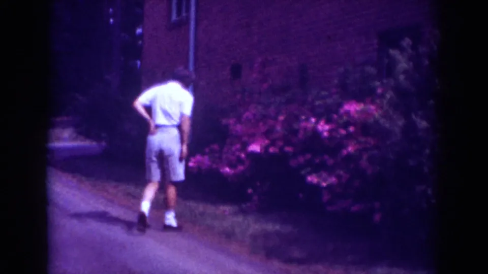 In Raleigh, 1962, a man in a white shirt and light shorts walks beside flowering bushes near a brick building.