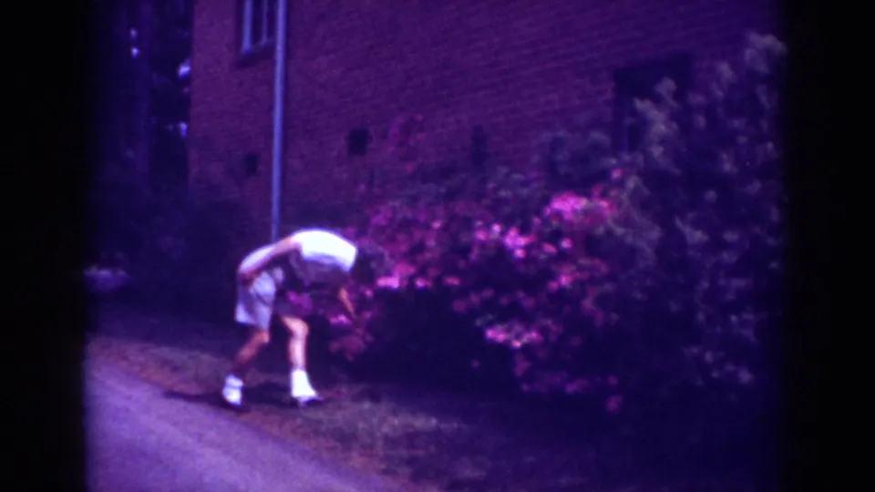 Raleigh 1962 home-movie frame showing a man bending to pick something up near flowering shrubs by a brick wall.