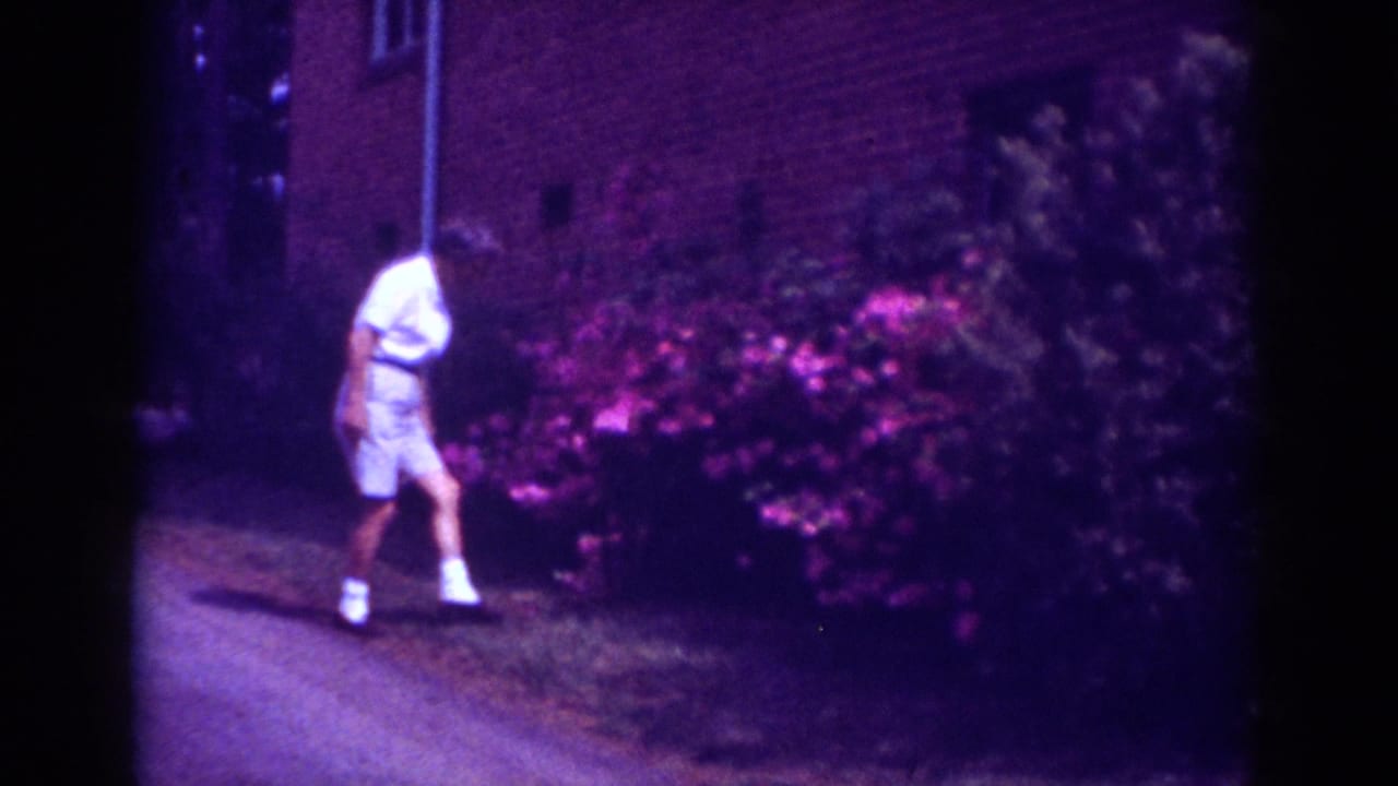 In 1962 Raleigh home-movie footage, a boy walks along a path beside bright pink garden shrubs and a brick exterior.