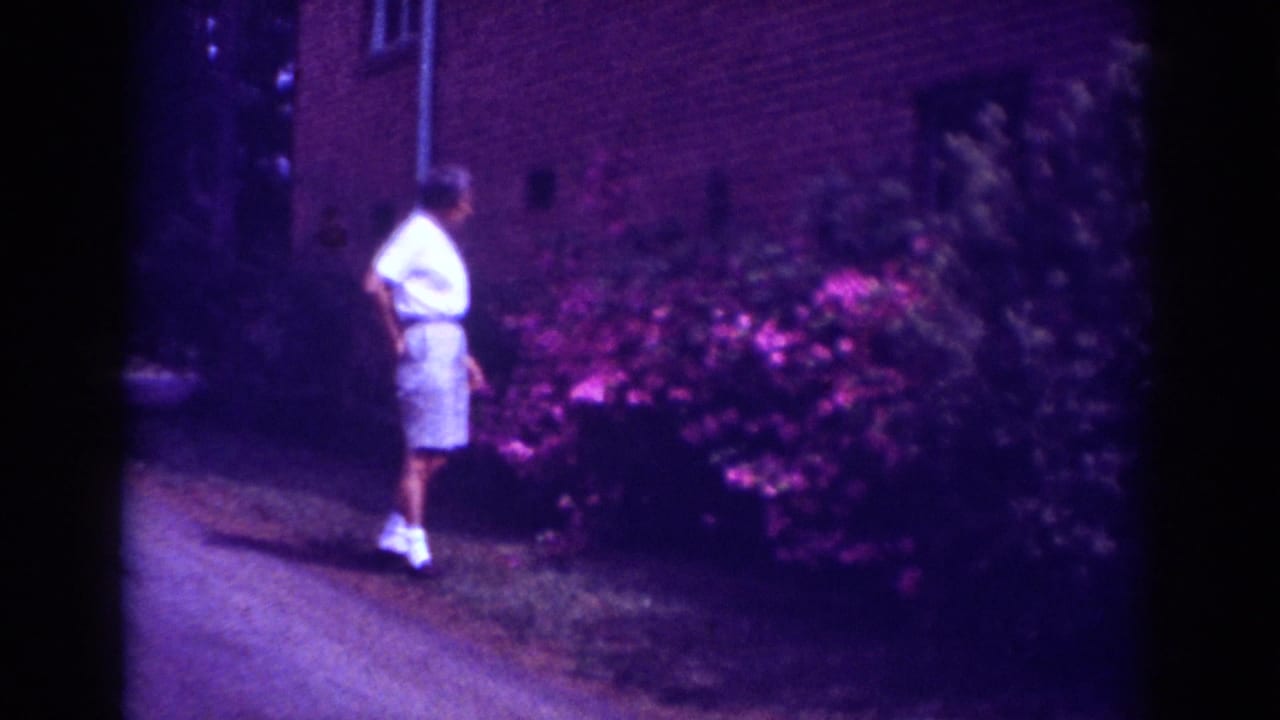 A boy in light summer clothing bends toward a flowering shrub outside a brick building in Raleigh home-movie footage.