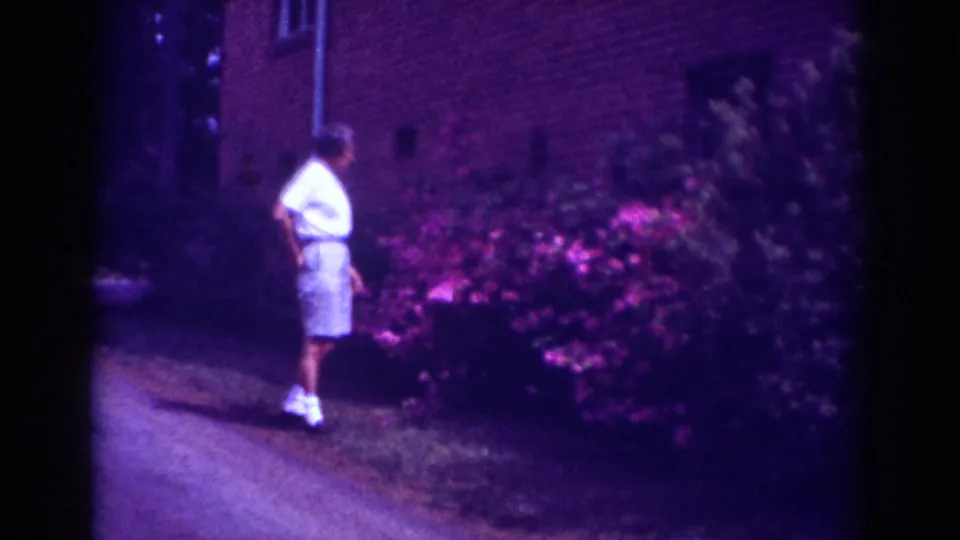 A boy in light summer clothing bends toward a flowering shrub outside a brick building in Raleigh home-movie footage.