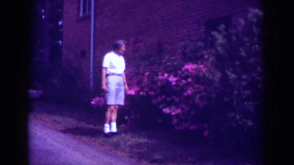 The boy pauses beside the pink shrubs, with a brick wall, shaded path, and soft 8mm color shift visible around him.