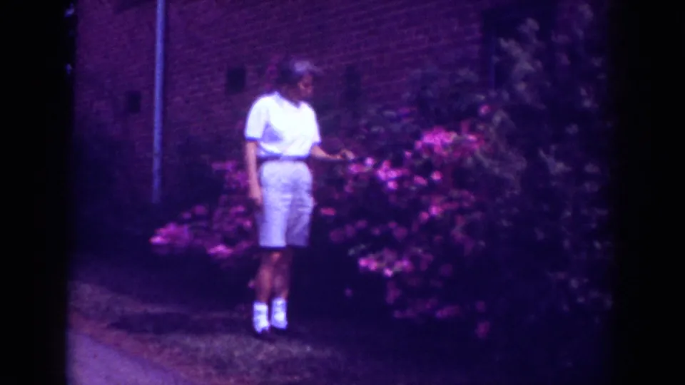 A boy stands close to the pink flowering bush and appears to gesture toward the blossoms outside a brick building.