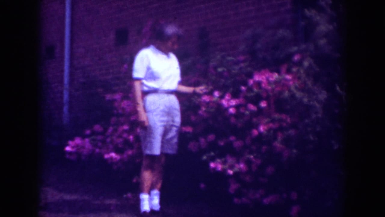 1962 Raleigh home-movie frame of a woman standing near pink flowering bushes against a brick wall.