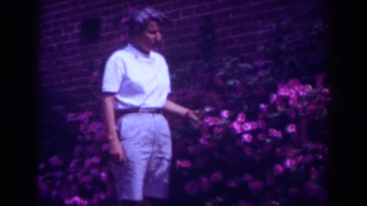 Adult woman in light summer clothing touches pink flowers in a Raleigh garden scene from 1962 home-movie footage.