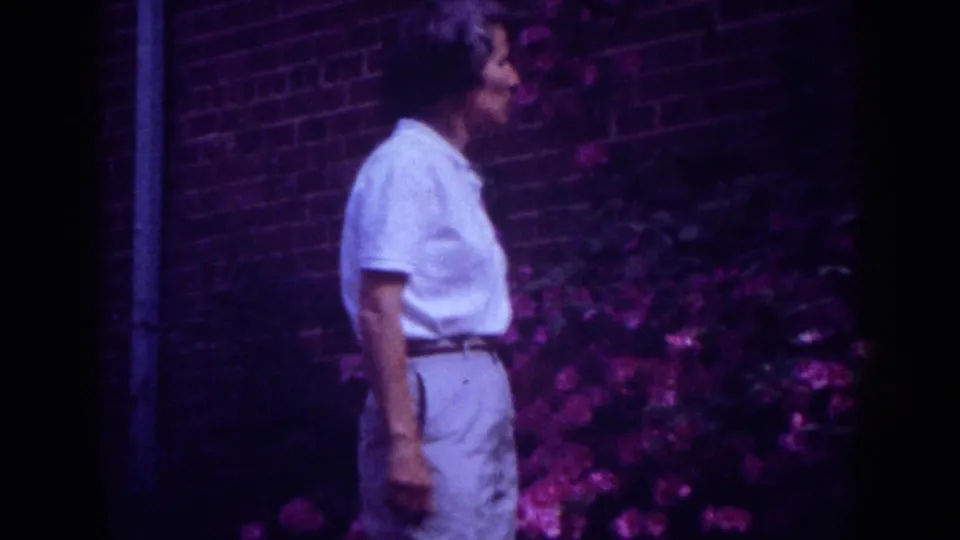 Side-profile view of the woman standing by pink blossoms in the 1962 Raleigh garden footage.