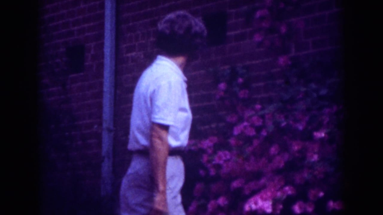 In 1962 Raleigh home-movie footage, an adult woman in a white short-sleeved shirt stands near pink flowering shrubs against a brick wall.