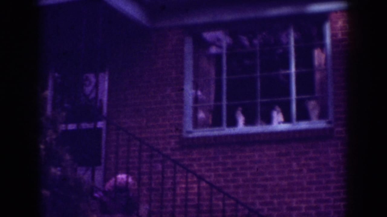 1962 Raleigh home-movie frame showing a brick house exterior at night, with a window, porch railing, and a person near the entry.