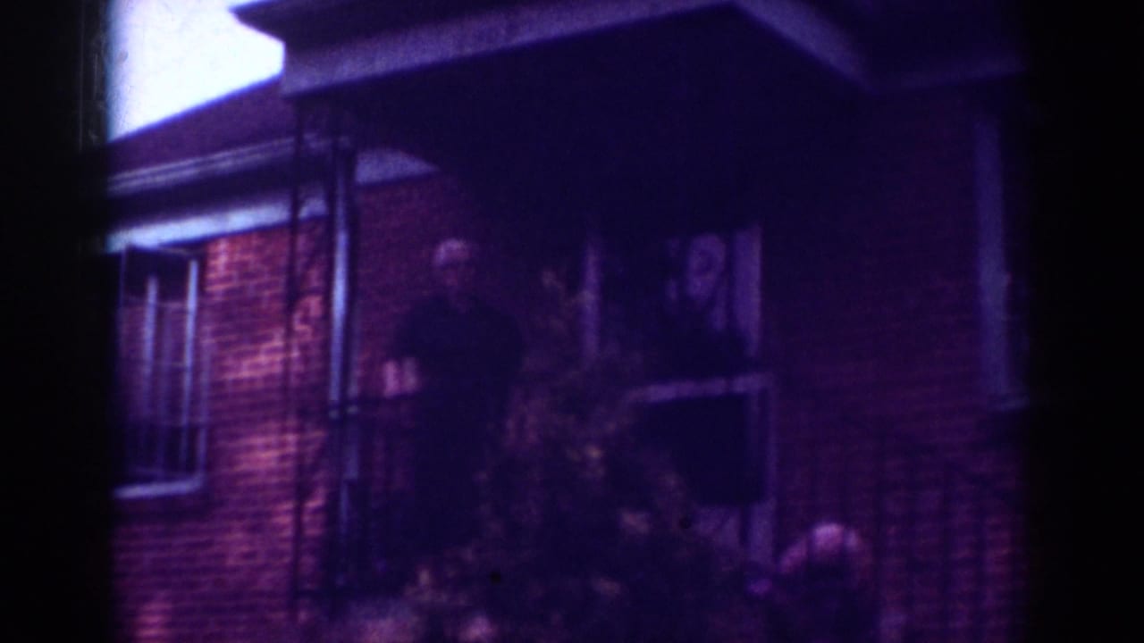 1962 Raleigh frame of a man in dark clothing standing by a brick house entrance at night, with porch details partly visible.